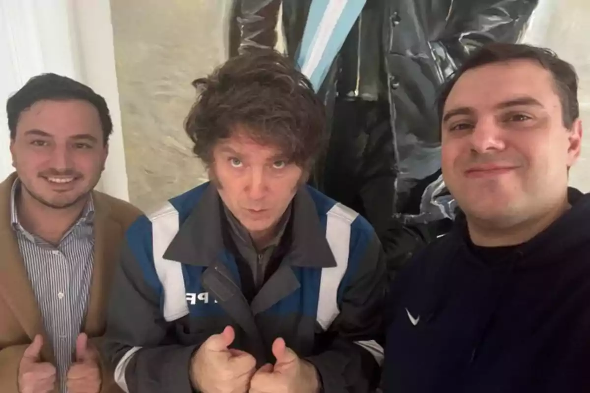 Tres hombres posan para una selfie sonriendo y haciendo el gesto de pulgares arriba frente a un cuadro